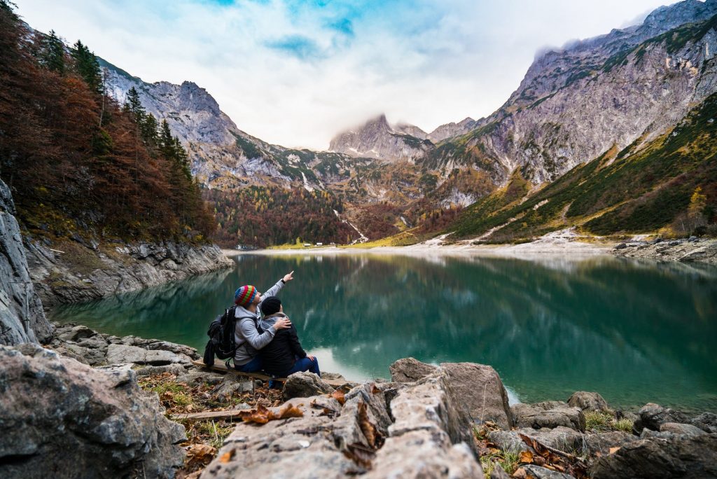 Um casal em um lindo visual cercado por montanhas e um lago abaixo, estão olhando e apontando para o Céu. O esquecimento de nossa origem Cósmica.
