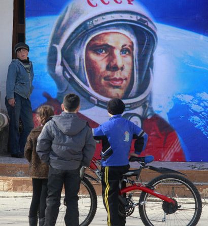 Um grande quadro com a imagem de Yuri Gagarin e três homens à frente um com bicicleta admirando o quadro. Planetário da Gávea comemora 50 anos da chegada do homem ao espaço.