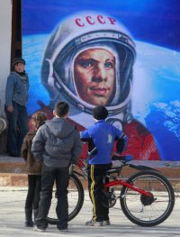 Um grande quadro com a imagem de Yuri Gagarin e três homens à frente um com bicicleta admirando o quadro. Planetário da Gávea comemora 50 anos da chegada do homem ao espaço.